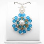 Large handmade statement pendant featuring multiple layers of large and small faceted aquamarine crystals surrounding an iridescent golden centre crystal, accented with white pearl beads and silver-tone seed beads, with a white pearl and silver-tone decorative bail, photographed on a white display stand.