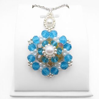 Large handmade statement pendant featuring multiple layers of large and small faceted aquamarine crystals surrounding an iridescent golden centre crystal, accented with white pearl beads and silver-tone seed beads, with a white pearl and silver-tone decorative bail, photographed on a white display stand.