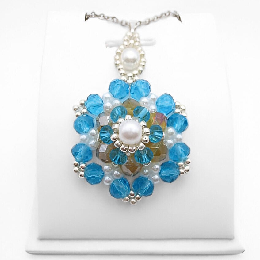Large handmade statement pendant featuring multiple layers of large and small faceted aquamarine crystals surrounding an iridescent golden centre crystal, accented with white pearl beads and silver-tone seed beads, with a white pearl and silver-tone decorative bail, photographed on a white display stand.
