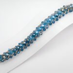 Handmade wide teal blue AB crystal bracelet featuring large iridescent faceted crystals and smaller blue crystals woven with silver seed bead accents in a full cluster pattern, displayed flat on a white jewellery stand showing full length and silver toggle clasp against a white background.