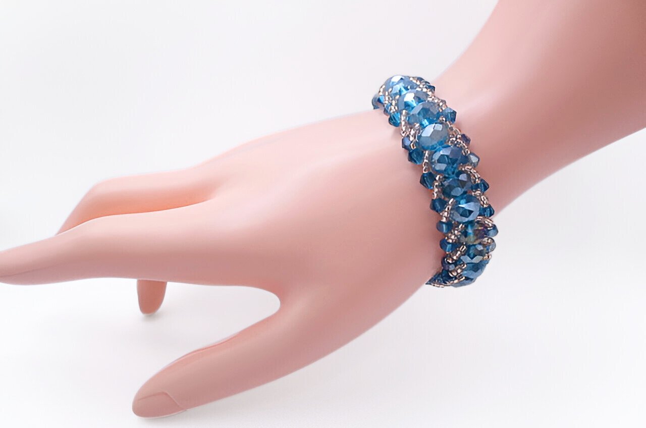 Handmade wide bracelet featuring large teal blue AB iridescent faceted crystals and smaller matching blue crystals woven together with silver seed bead accents in a lush cluster design, shown worn on a mannequin wrist against a white background.