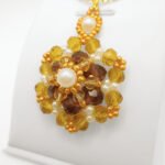 Side view of the statement pendant featuring rich amber and golden faceted crystals, a white pearl centre, white seed pearl accents and gold seed bead detailing throughout, with a decorative pearl and gold bail, displayed on a gold-tone chain and white jewellery stand.