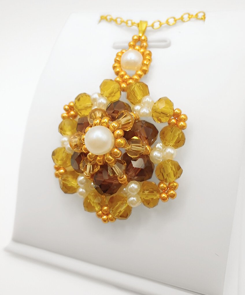 Side view of the statement pendant featuring rich amber and golden faceted crystals, a white pearl centre, white seed pearl accents and gold seed bead detailing throughout, with a decorative pearl and gold bail, displayed on a gold-tone chain and white jewellery stand.
