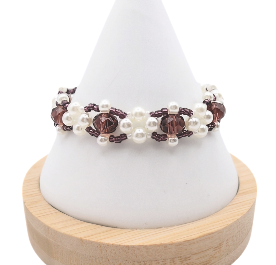 Handmade beaded bracelet featuring faceted deep purple crystal beads surrounded by clusters of white pearl beads and small purple seed bead accents, displayed on a white cone jewelry stand with wooden base.