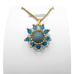 Flower-shaped aquamarine pendant with a smooth round aquamarine centre stone surrounded by alternating teal blue and clear faceted crystals, accented with gold and silver-tone beads, displayed on a gold-tone curb chain and white jewellery stand.