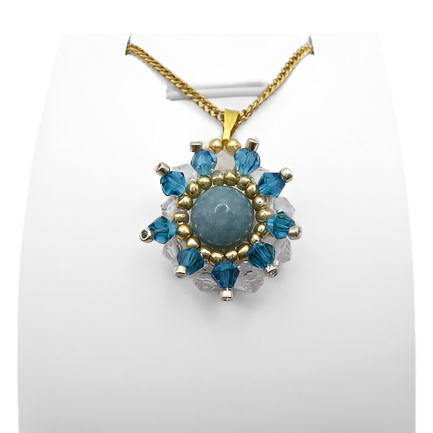aquamarine-pendant-display Flower-shaped aquamarine pendant with a smooth round aquamarine centre stone surrounded by alternating teal blue and clear faceted crystals, accented with gold and silver-tone beads, displayed on a gold-tone curb chain and white jewellery stand.