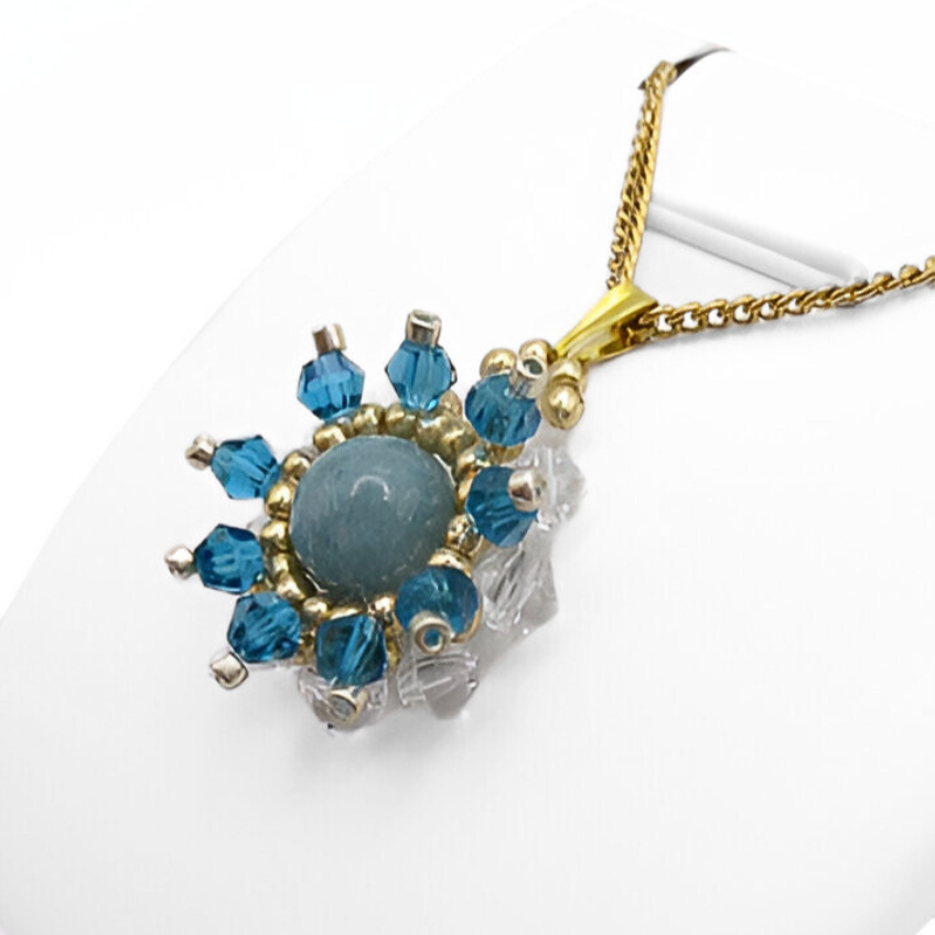 aquamarine-pendant-side-view Side view of the aquamarine pendant with a smooth round aquamarine centre stone surrounded by alternating teal blue and clear faceted crystals, accented with gold and silver-tone beads, displayed on a gold-tone curb chain and white jewellery stand.