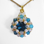 Blue crystal pendant necklace featuring a large teal blue faceted centre stone surrounded by white pearls, aquamarine and clear crystals, with a gold-tone bail and a gold-tone chain, photographed on a white display stand.