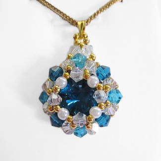 Blue crystal pendant necklace featuring a large teal blue faceted centre stone surrounded by white pearls, aquamarine and clear crystals, with a gold-tone bail and a gold-tone chain, photographed on a white display stand.