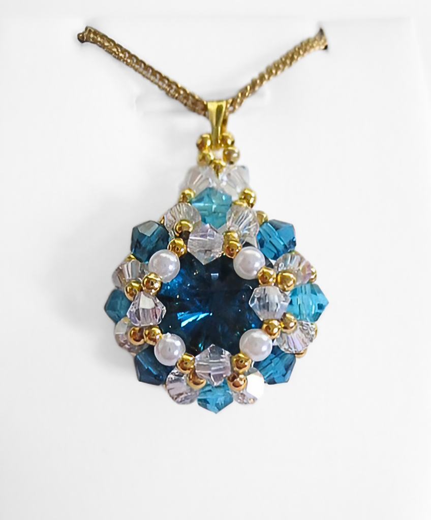 blue-crystal-pendant-closeup Blue crystal pendant necklace featuring a large teal blue faceted centre stone surrounded by white pearls, aquamarine and clear crystals, with a gold-tone bail and a gold-tone chain, photographed on a white display stand.