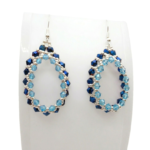 Handmade beaded teardrop hoop earrings featuring metallic sapphire blue and aqua light blue crystals with silver seed beads on silver-tone ear wires, displayed on a white jewellery stand.