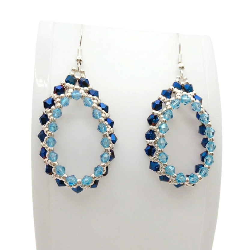 blue-teardrop-hoop-earrings-display Handmade beaded teardrop hoop earrings featuring metallic sapphire blue and aqua light blue crystals with silver seed beads on silver-tone ear wires, displayed on a white jewellery stand.