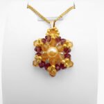 Crystal and pearl pendant with a champagne centre pearl surrounded by warm champagne faceted crystals and an outer purple crystals, accented throughout with gold beads, displayed on a gold-tone curb chain and white jewellery stand.