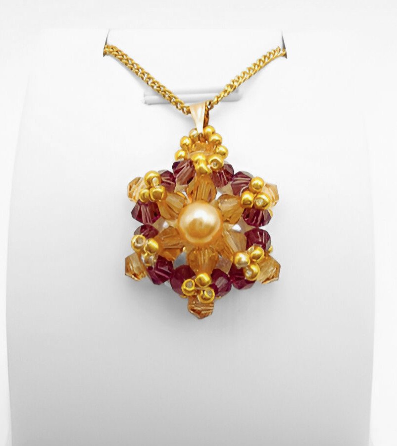 champange-pearl crystal-pendant-display Crystal and pearl pendant with a champagne centre pearl surrounded by warm champagne faceted crystals and an outer purple crystals, accented throughout with gold beads, displayed on a gold-tone curb chain and white jewellery stand.