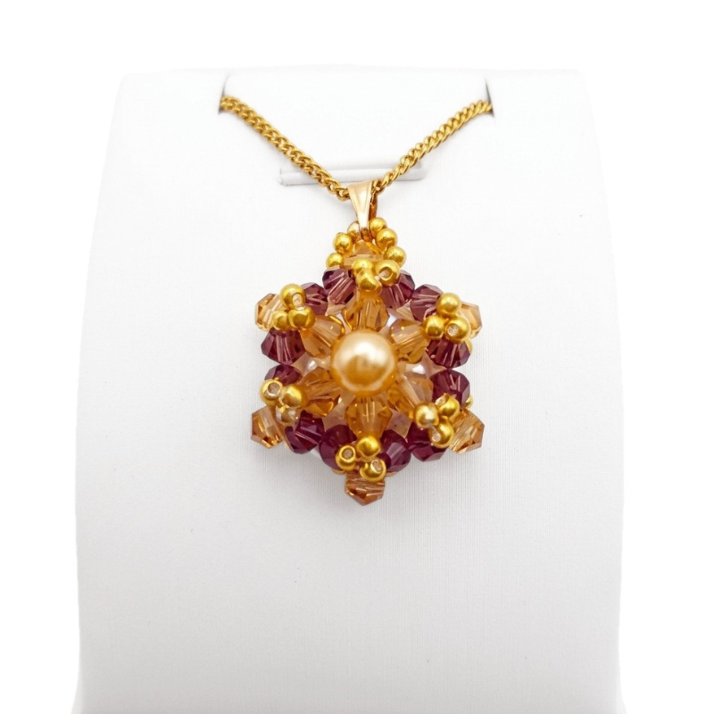 Crystal and pearl pendant with a champagne centre pearl surrounded by warm champagne faceted crystals and an outer purple crystals, accented throughout with gold beads, displayed on a gold-tone curb chain and white jewellery stand.