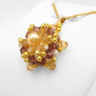 Side view of the crystal and pearl pendant with a champagne centre pearl surrounded by golden champagne crystals and an outer purple crystals, accented throughout with gold beads, displayed on a gold-tone curb chain and white jewellery stand.