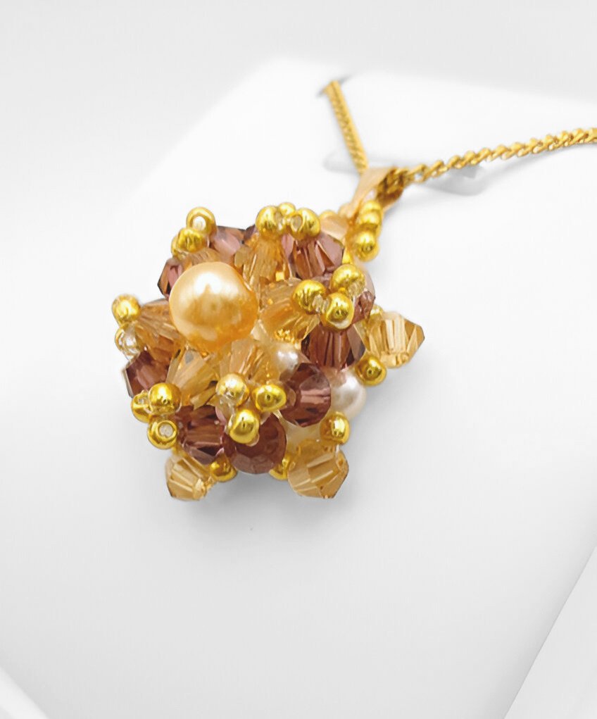 champange-pearl-pendant-side-view Side view of the crystal and pearl pendant with a champagne centre pearl surrounded by golden champagne crystals and an outer purple crystals, accented throughout with gold beads, displayed on a gold-tone curb chain and white jewellery stand.