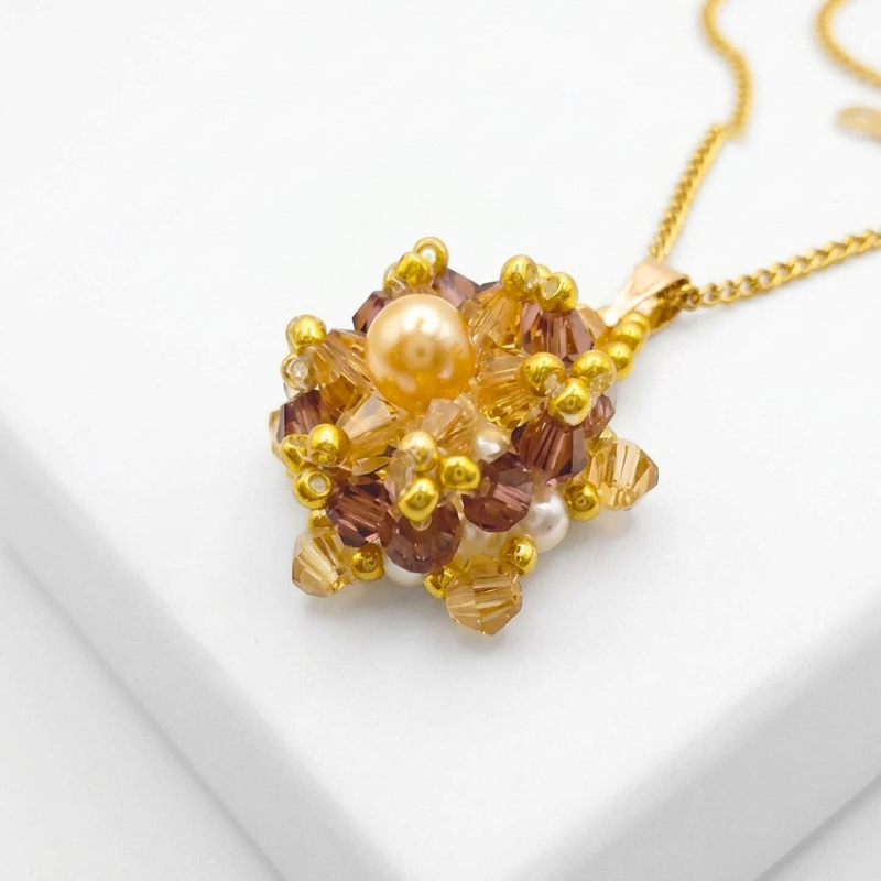 Angled view of the crystal and pearl pendant with a champagne centre pearl surrounded by golden champagne crystals and an outer purple crystals, accented throughout with gold beads, displayed on a gold-tone curb chain and white jewellery stand.