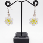 Close-up of handmade beaded daisy earrings showing intricate white seed bead petals, pearl accent beads, and a bright yellow seed bead flower centre, with silver hook findings on a dark background.