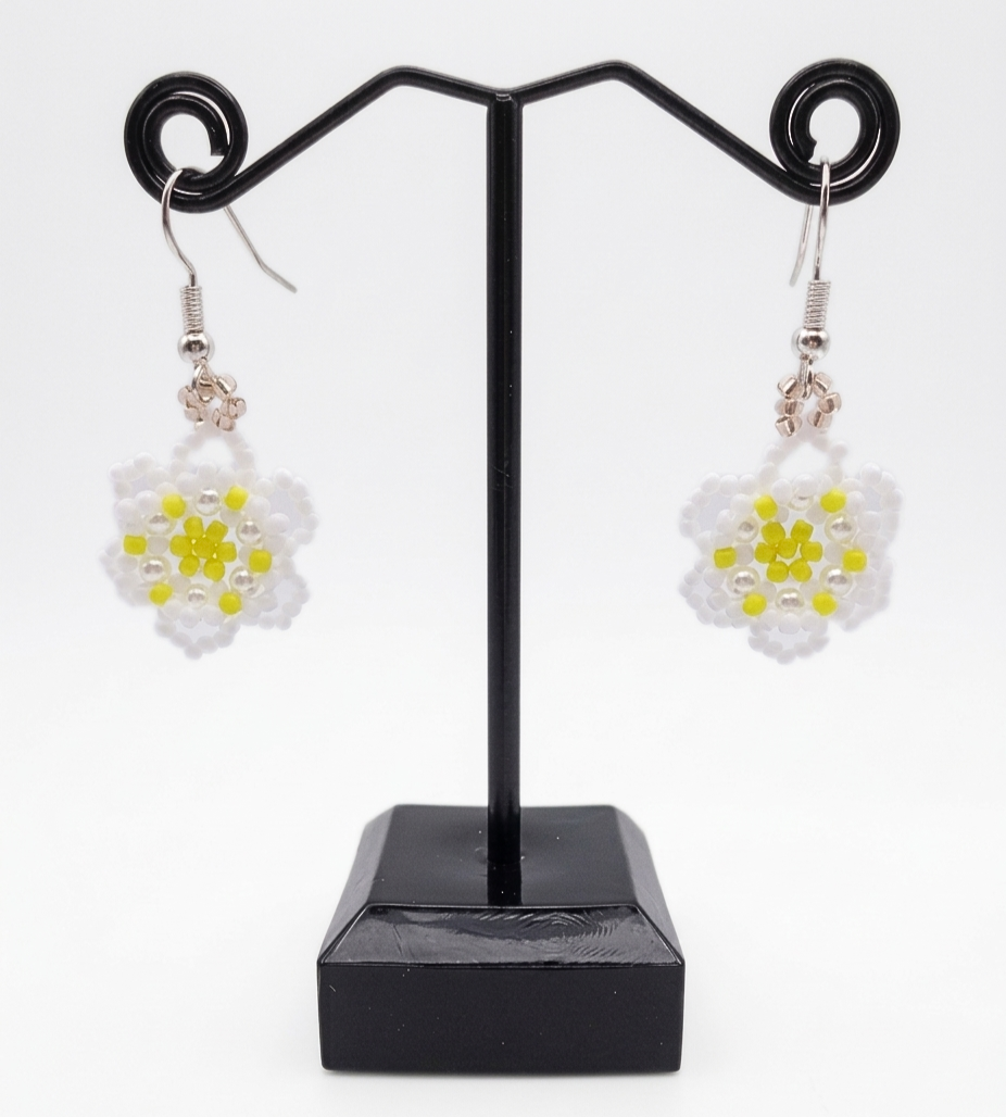 floral-earrings-display Close-up of handmade beaded daisy earrings showing intricate white seed bead petals, pearl accent beads, and a bright yellow seed bead flower centre, with silver hook findings on a dark background.