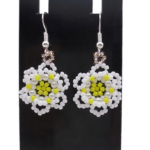 A pair of handmade white and yellow beaded daisy flower earrings hanging on a black metal earring display stand against a white background.