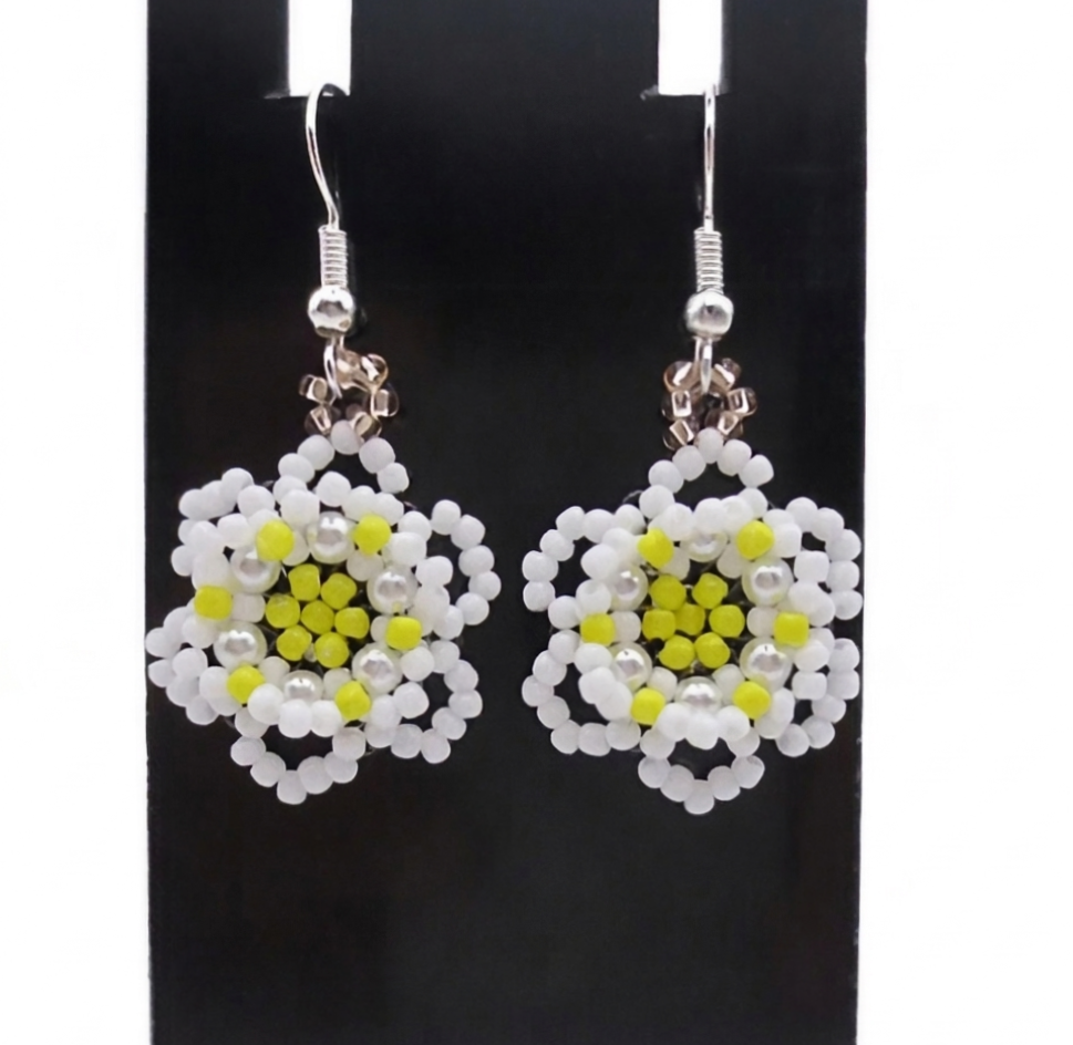floral-earrings-flat-display A pair of handmade white and yellow beaded daisy flower earrings hanging on a black metal earring display stand against a white background.