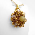 Side angle view of a handmade golden tourmaline pendant featuring a smooth round yellow tourmaline centre stone surrounded by warm amber and champagne faceted crystals with gold seed bead accents, displayed on a gold-tone chain and white jewellery stand.