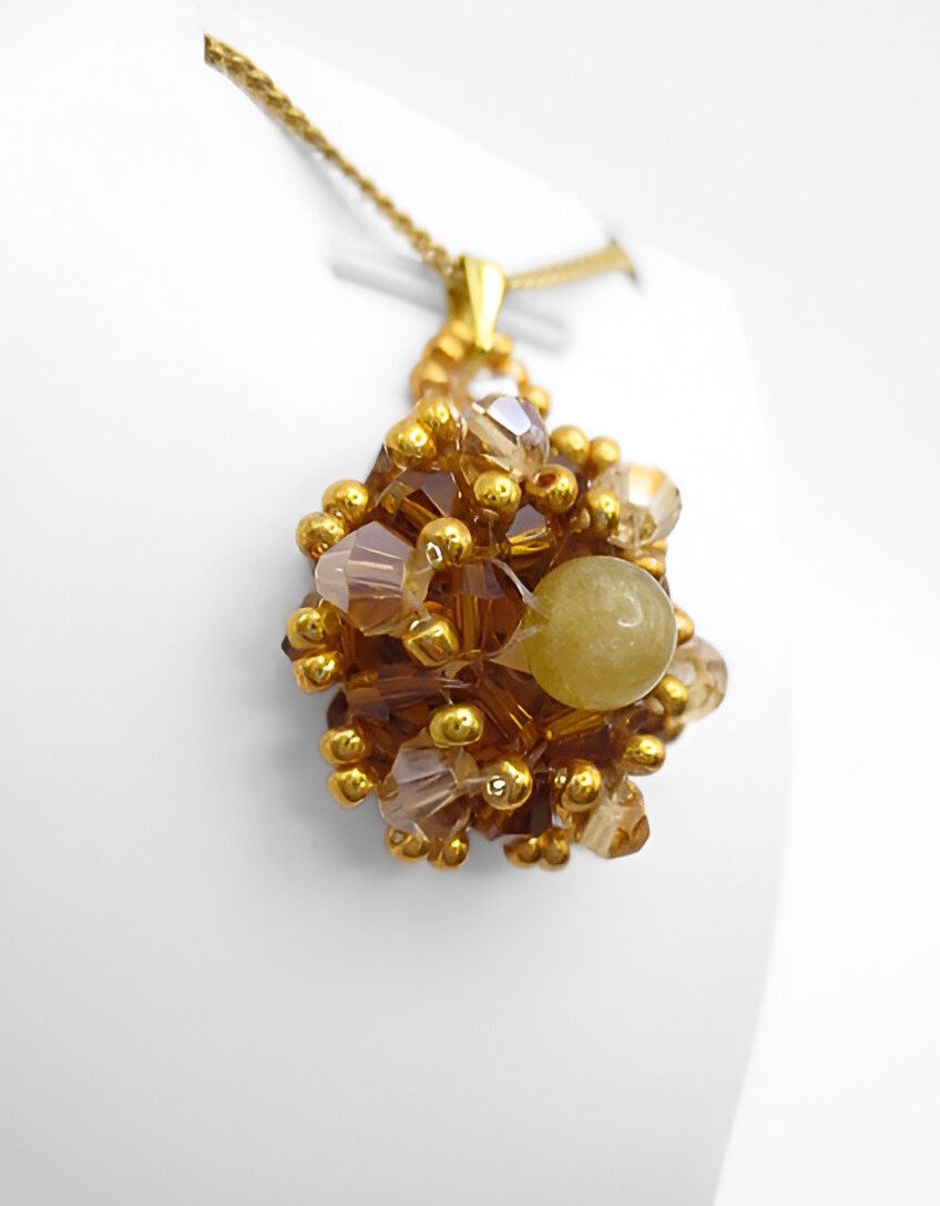 golden-tourmaline- pendant-display-side Side angle view of a handmade golden tourmaline pendant featuring a smooth round yellow tourmaline centre stone surrounded by warm amber and champagne faceted crystals with gold seed bead accents, displayed on a gold-tone chain and white jewellery stand.