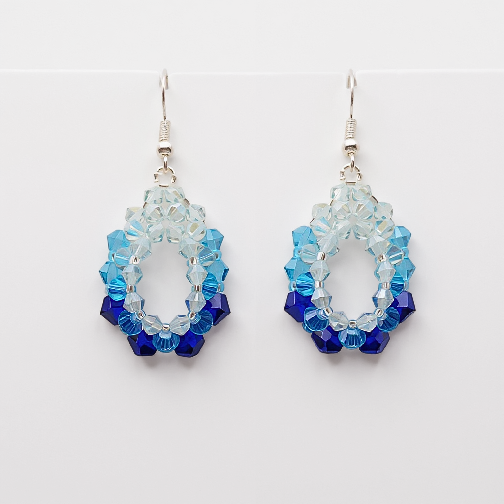 Handmade blue ombre crystal oval hoop dangle earrings, gradient sapphire to sky blue beaded open frame, silver ear wire, shown against a plain white background