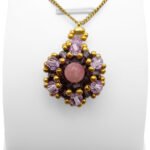 Handmade pink tourmaline pendant featuring a smooth round pink tourmaline centre stone surrounded by deep violet and light purple faceted crystals with gold bead accents and a gold-tone bail, shown on a white jewellery stand.