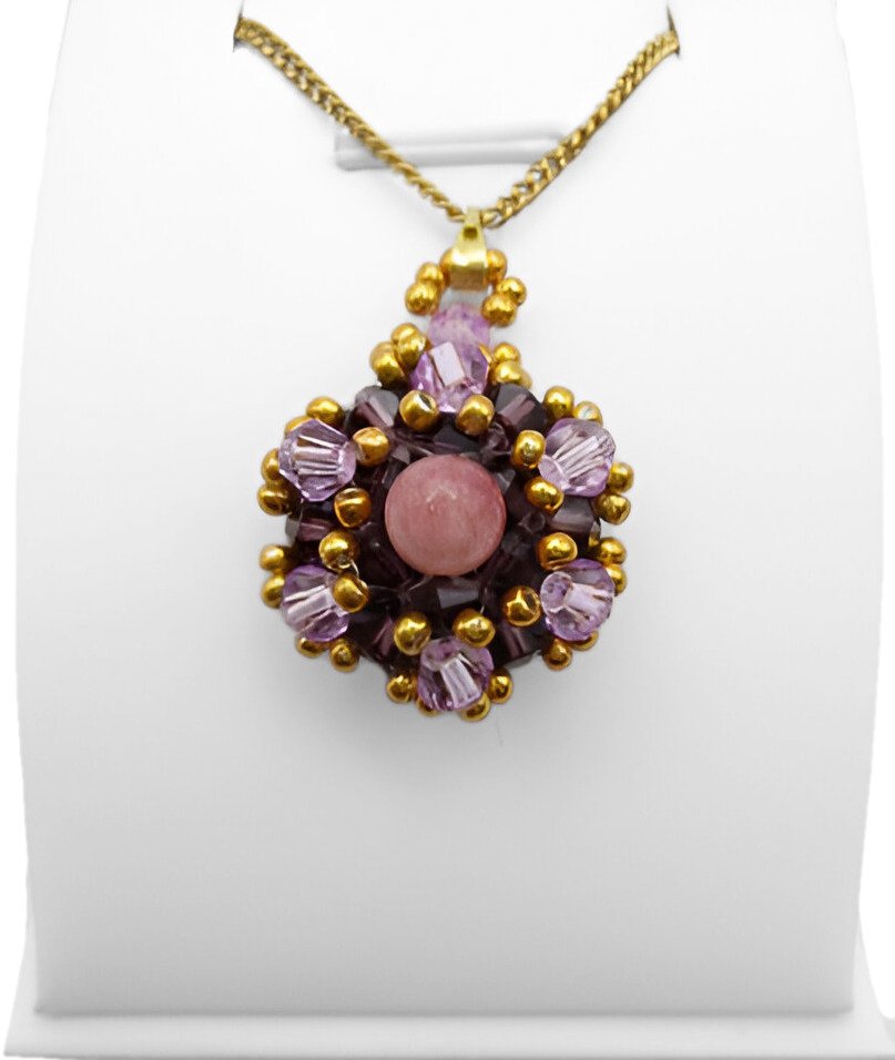 pink-tourmaline pendant-display Handmade pink tourmaline pendant featuring a smooth round pink tourmaline centre stone surrounded by deep violet and light purple faceted crystals with gold bead accents and a gold-tone bail, shown on a white jewellery stand.