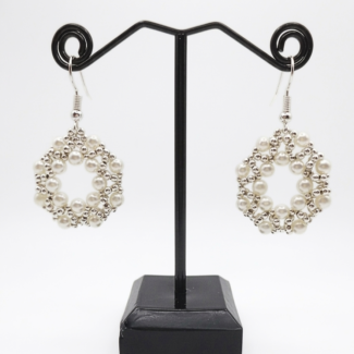Handmade white faux pearl and silver-tone beaded open hoop drop earrings displayed on a black metal earring stand, showing the full shape and drape of the woven circular lattice design