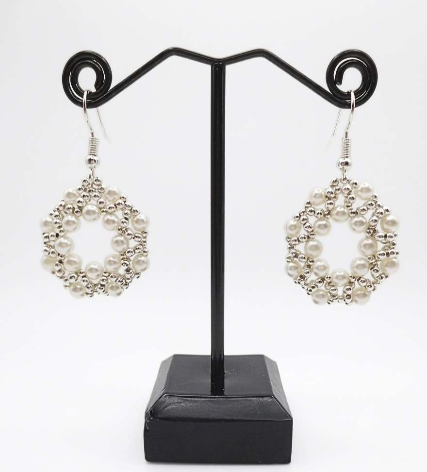 Handmade white faux pearl and silver-tone beaded open hoop drop earrings displayed on a black metal earring stand, showing the full shape and drape of the woven circular lattice design