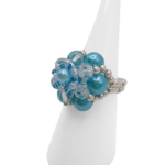 A side view of a handmade aquamarine pearl and crystal cluster ring on white cone ring stand — sky blue pearl beads, clear crystals, and silver-tone seed bead band