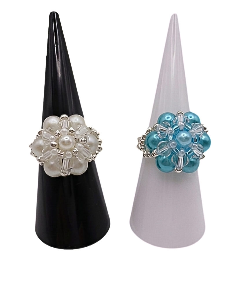 Two handmade pearl cluster cocktail rings displayed side by side on black and white cone stands — white pearl ring on left, aquamarine blue pearl ring on right, both with clear crystal and silver-tone accents
