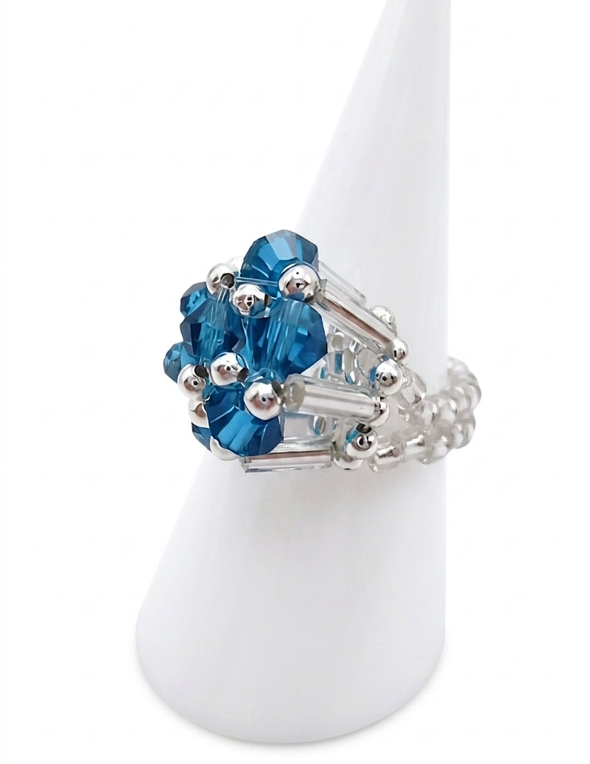Angled side view of handmade ocean blue and clear crystal cluster ring on white cone display stand — faceted ocean blue crystals, clear rod crystals, and silver-tone beaded band