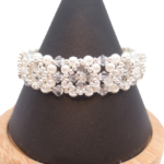 Bridal bracelet featuring a repeating floral pattern of white pearls and clear faceted crystals with silver-tone seed bead accents throughout, displayed on a dark cone bracelet stand with a wooden base against a white background.