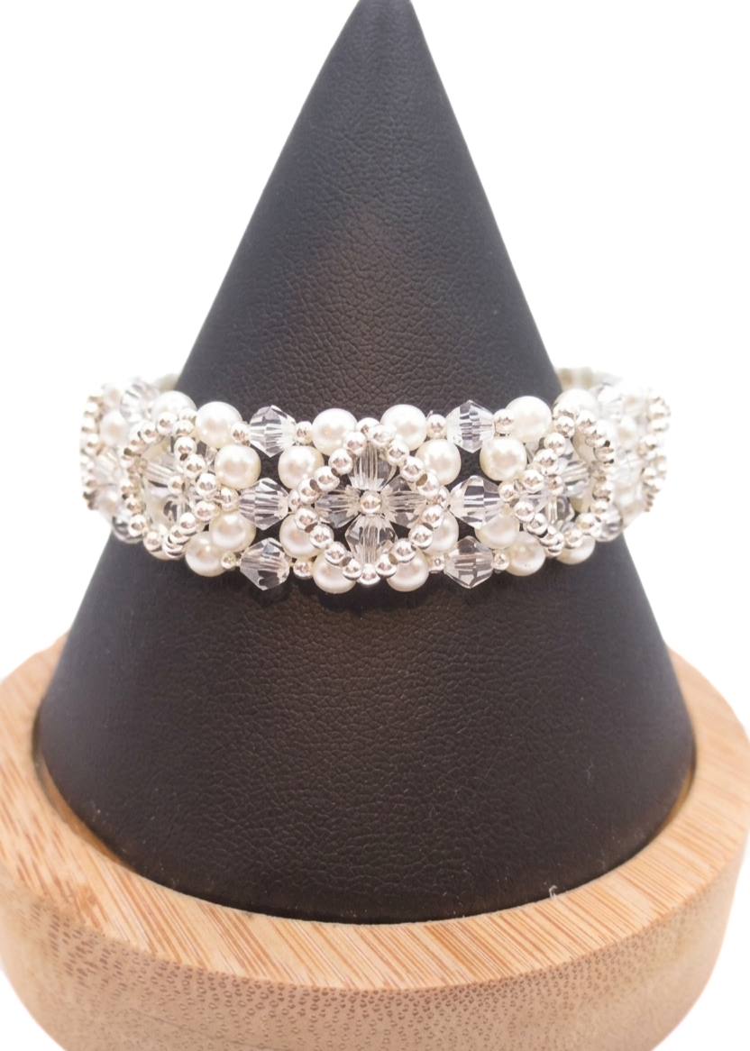 Bridal bracelet featuring a repeating floral pattern of white pearls and clear faceted crystals with silver-tone seed bead accents throughout, displayed on a dark cone bracelet stand with a wooden base against a white background.