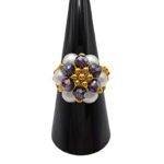 Front view of a handmade floral cluster ring with white pearl beads, faceted purple AB iridescent crystal beads, and gold-tone metal accents, displayed on a black ring cone