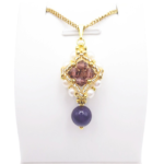 Handmade purple drop pendant necklace featuring a diamond-shaped top section with purple faceted crystals, white pearls and gold beads, suspending a smooth violet gemstone drop bead, displayed on a gold-tone curb chain and white jewellery stand.