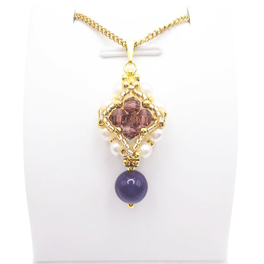 Handmade purple drop pendant necklace featuring a diamond-shaped top section with purple faceted crystals, white pearls and gold beads, suspending a smooth violet gemstone drop bead, displayed on a gold-tone curb chain and white jewellery stand.