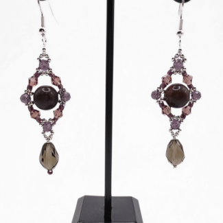Handmade drop earrings with a dark tourmaline stone centrepiece, open diamond-shaped silver frame adorned with purple and mauve crystals, and a dangling faceted smoky grey crystal teardrop, displayed on a black metal earring stand.