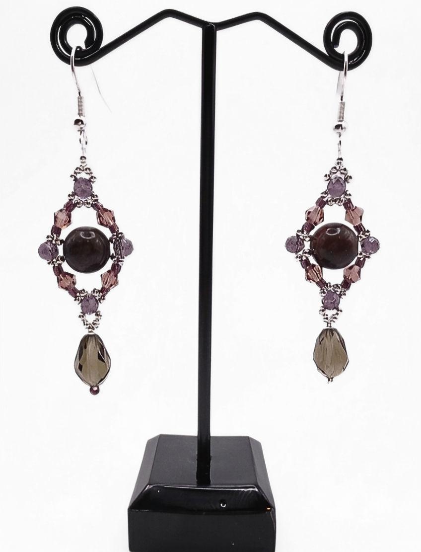 Handmade drop earrings with a dark tourmaline stone centrepiece, open diamond-shaped silver frame adorned with purple and mauve crystals, and a dangling faceted smoky grey crystal teardrop, displayed on a black metal earring stand.