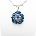 Close-up view of floral pendant featuring white pearls and a blue pearl centre accent surrounded by metallic blue faceted crystals and aquamarine bead detailing, with a silver-tone bail, displayed on a white stand.