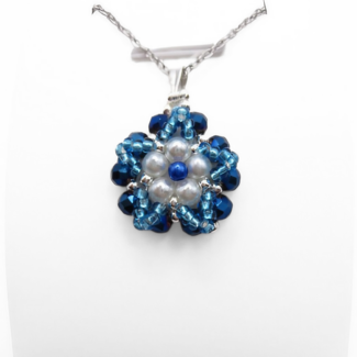 Close-up view of floral pendant featuring white pearls and a blue pearl centre accent surrounded by metallic blue faceted crystals and aquamarine bead detailing, with a silver-tone bail, displayed on a white stand.