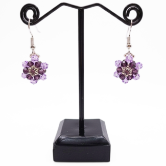 Lilac and purple crystal flower drop earrings hanging on a black metal jewellery stand against a white background