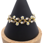 Handmade delicate daisy bracelet featuring white seed bead daisy flowers with yellow centres, white pearls, champagne crystals and gold seed bead accents in a repeating open floral chain design, displayed on a black cone bracelet stand with a wooden base against a white background.