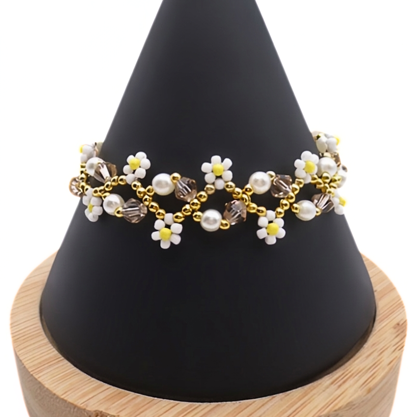 Handmade delicate daisy bracelet featuring white seed bead daisy flowers with yellow centres, white pearls, champagne crystals and gold seed bead accents in a repeating open floral chain design, displayed on a black cone bracelet stand with a wooden base against a white background.