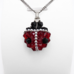 Ladybug-shaped pendant featuring vivid red and black faceted crystals with a silver bead spine and border detailing, black crystal head and a silver-tone bail, displayed on a white stand.