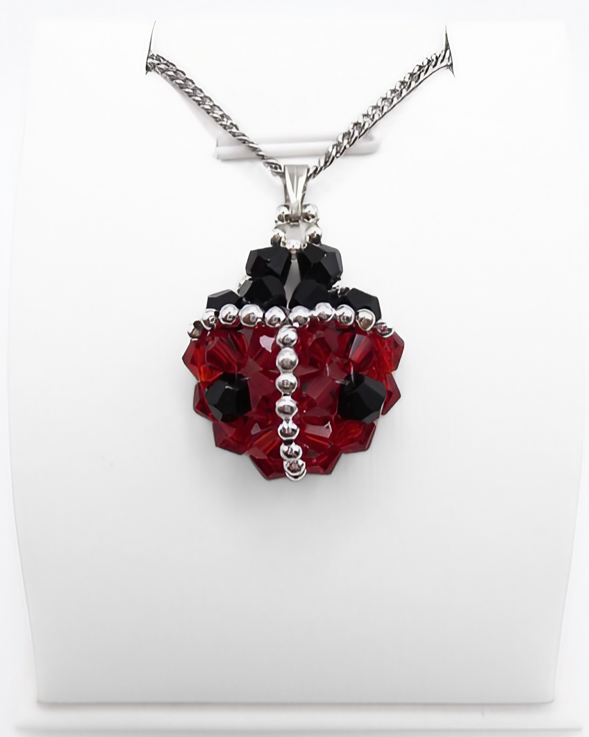Ladybug-shaped pendant featuring vivid red and black faceted crystals with a silver bead spine and border detailing, black crystal head and a silver-tone bail, displayed on a white stand.